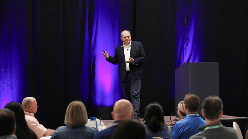 Richard Petti, CEO von IQGeo, präsentiert auf der Bühne des IQGeo Americas Meetup 2025 in Dallas vor einem vollen Publikum bei lila Bühnenbeleuchtung