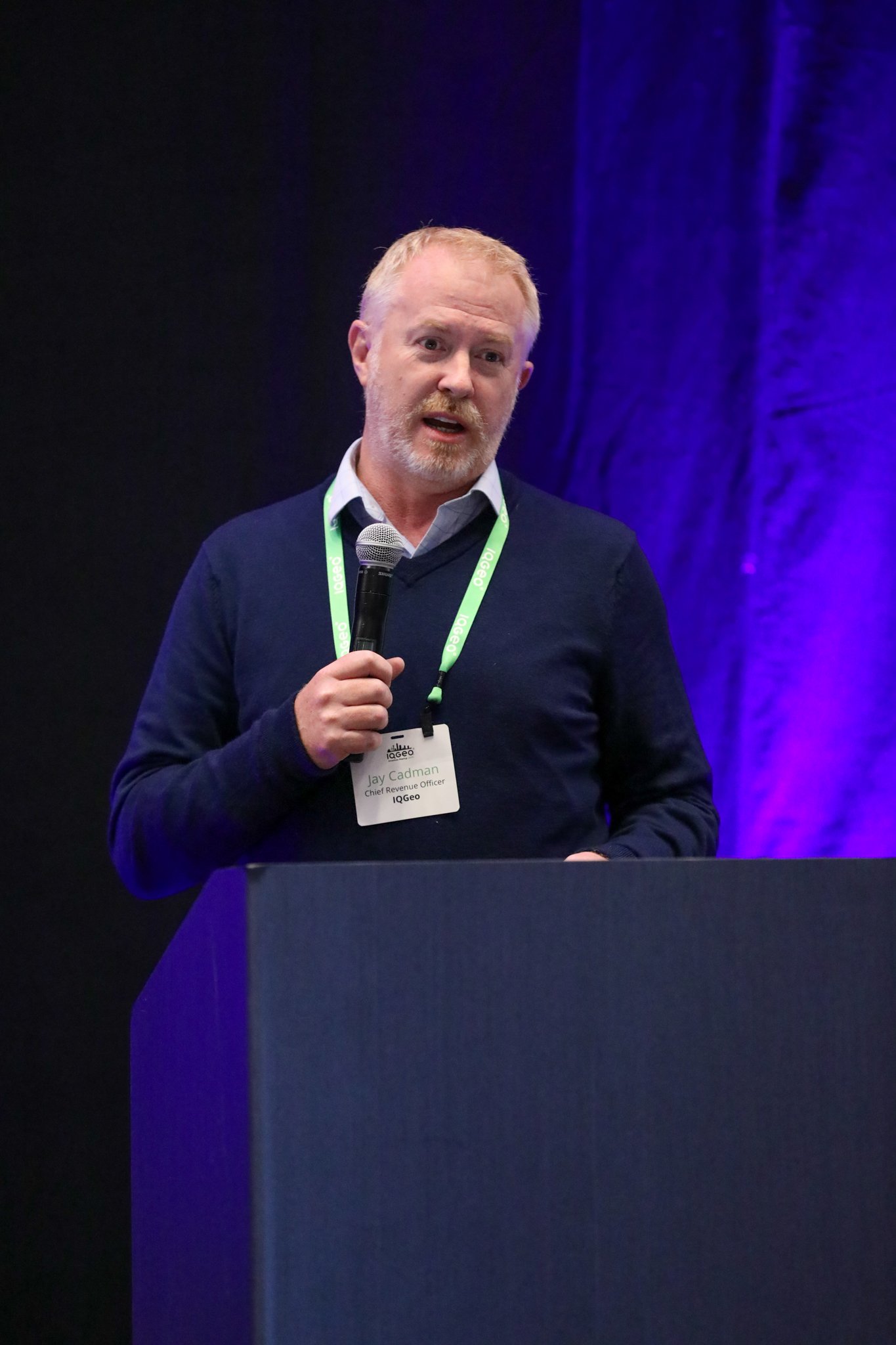 Jay Cadman von IQGeo präsentiert auf der Bühne beim IQGeo Americas Meetup 2025 in Dallas vor einem vollen Publikum bei lila Bühnenbeleuchtung