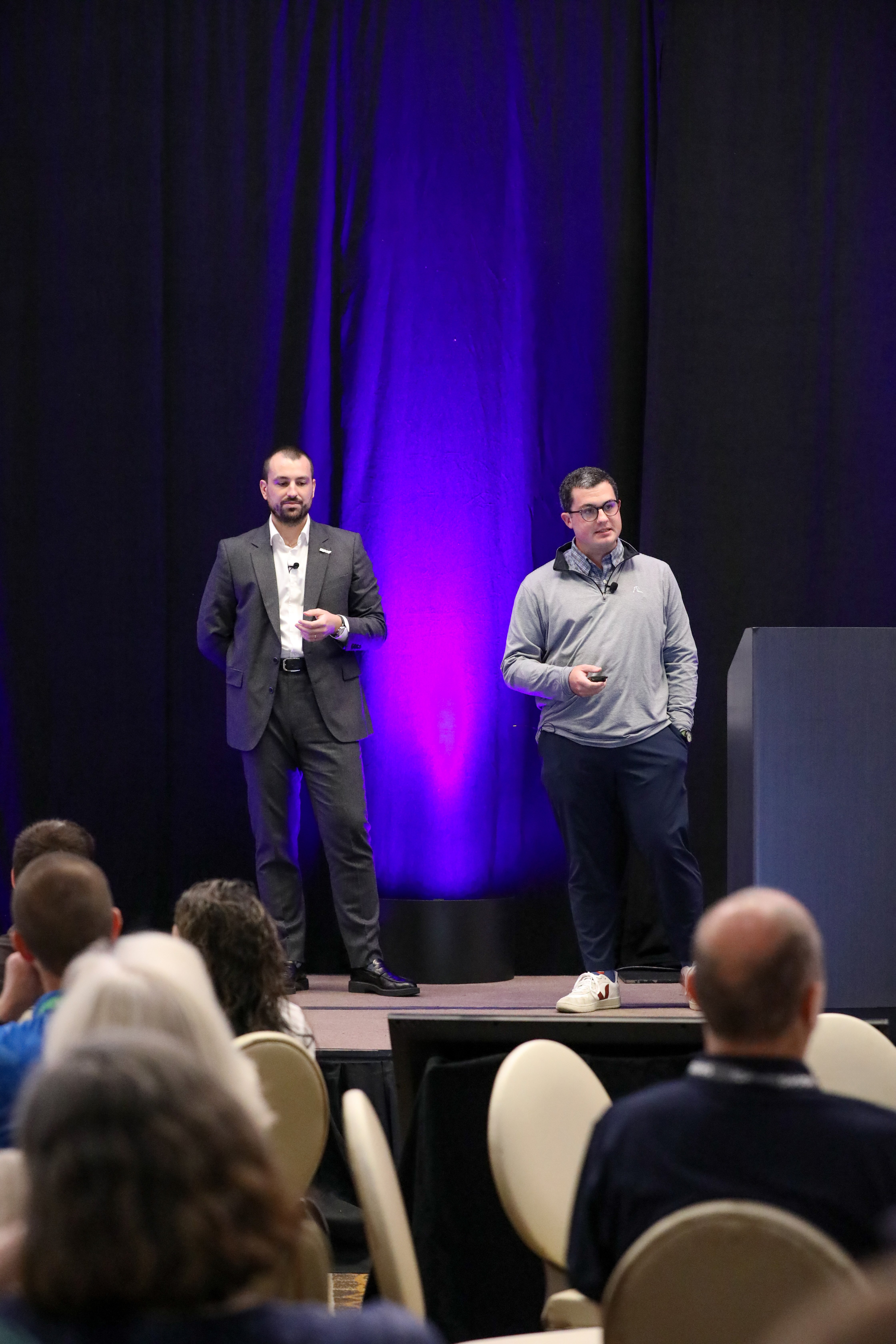Thomas Thuillier und Matt Roberts von IQGeo präsentieren auf der Bühne beim IQGeo Americas Meetup 2025 in Dallas vor einem vollen Publikum bei lila Bühnenbeleuchtung