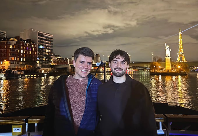 Zwei Menschen stehen nachts auf einem Uferweg, hinter ihnen leuchten die Gebäude der Stadt. Auf der anderen Seite des Flusses sind unter dem bewölkten Himmel ein beleuchteter Turm und eine Statue auf einer kleinen Insel zu sehen.