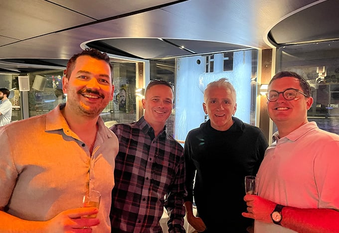 Vier Personen stehen bei einer gesellschaftlichen Veranstaltung zusammen in einem Innenraum, jeder hält ein Getränk in der Hand, im Hintergrund warme Beleuchtung und moderne Einrichtung.