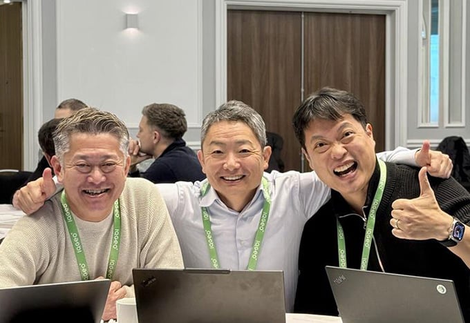 Drei Personen sitzen während einer Besprechung oder einem Workshop mit Laptops an einem Tisch, wobei eine Person den Daumen hochhält.