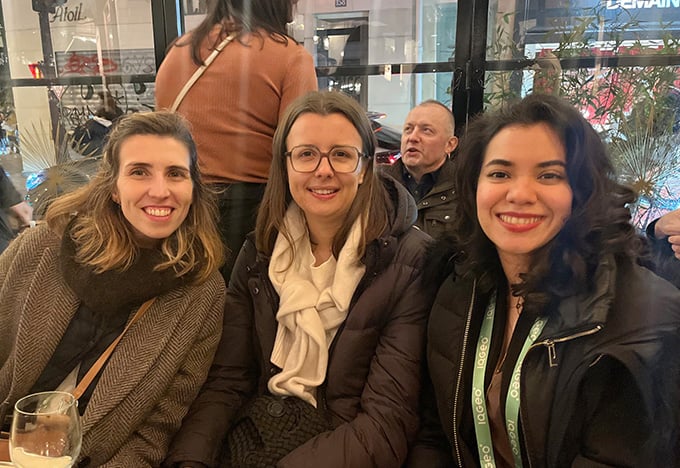 Drei Personen sitzen zusammen an einem Cafétisch, in Winterkleidung eingemummt, mit warmer Innenbeleuchtung und einem Blick auf eine Stadtstraße durch das Fenster hinter ihnen.