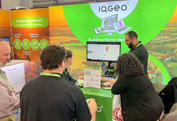  Menschen versammeln sich am Messestand von IQGeo, wo ein Moderator auf einem Monitor die Software vorführt.
