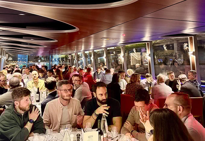 Eine große Gruppe von Menschen sitzt während eines lebhaften Abendessens auf einem Boot dicht beieinander an Tischen.